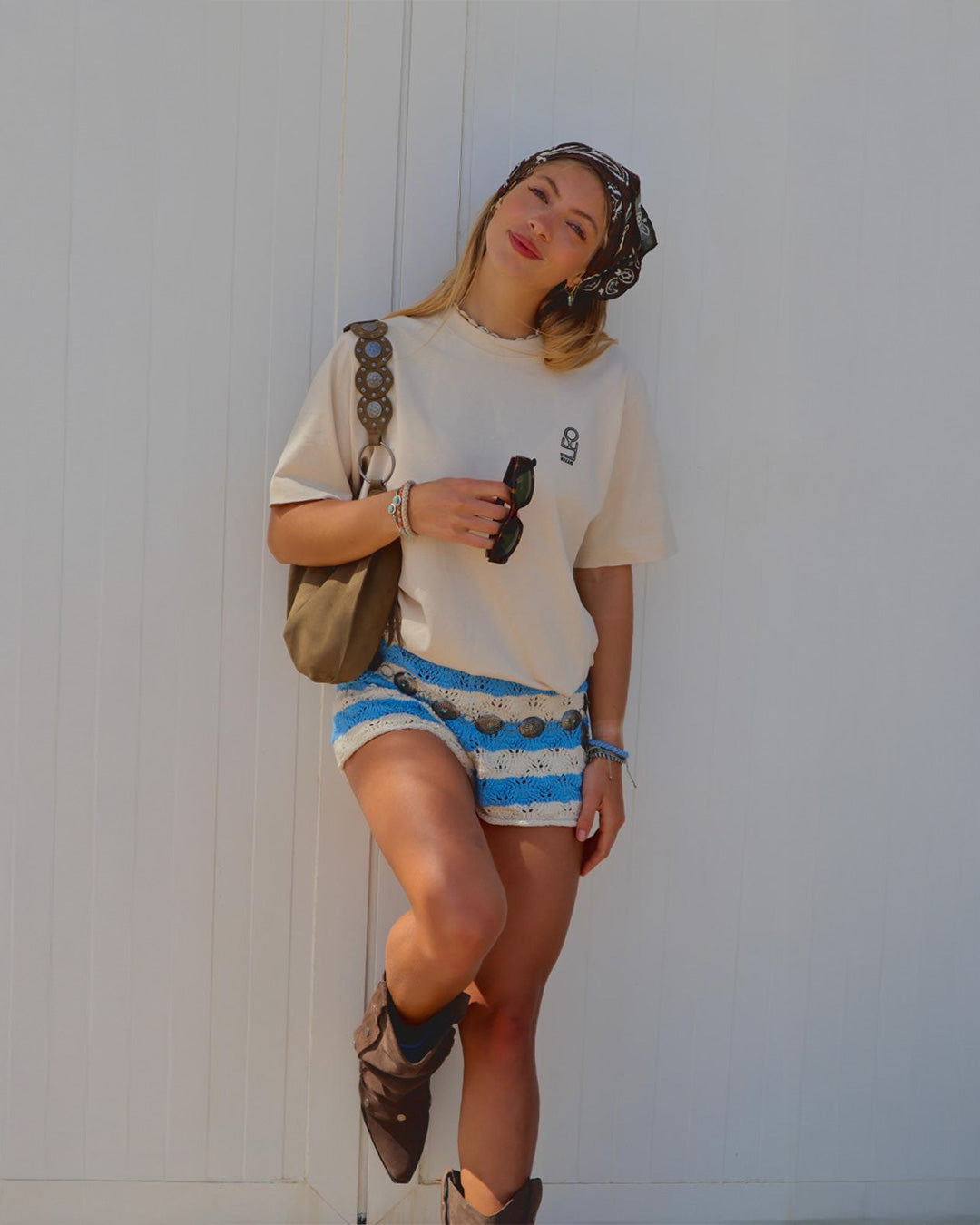 Jonge dame, blond haar, blank,  beige t-shirt, geborduurd logo, wit met blauwe broek, bruin tasje om schouder, bruine laarzen, witte houten muur.