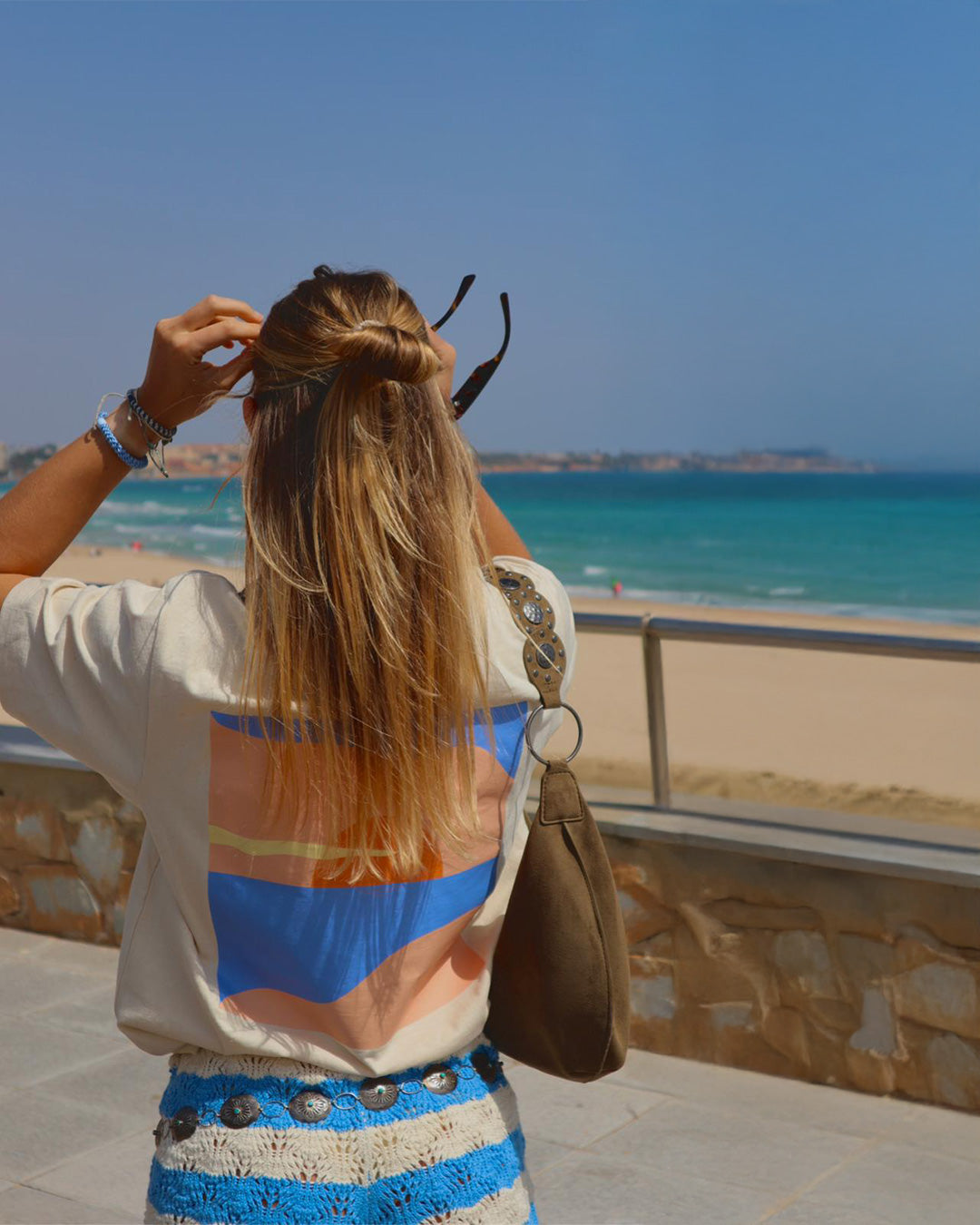 Blonde vrouw, blauw water, strand, beige t-shirt, print met pastel tonen op de rug, wit met blauwe broek