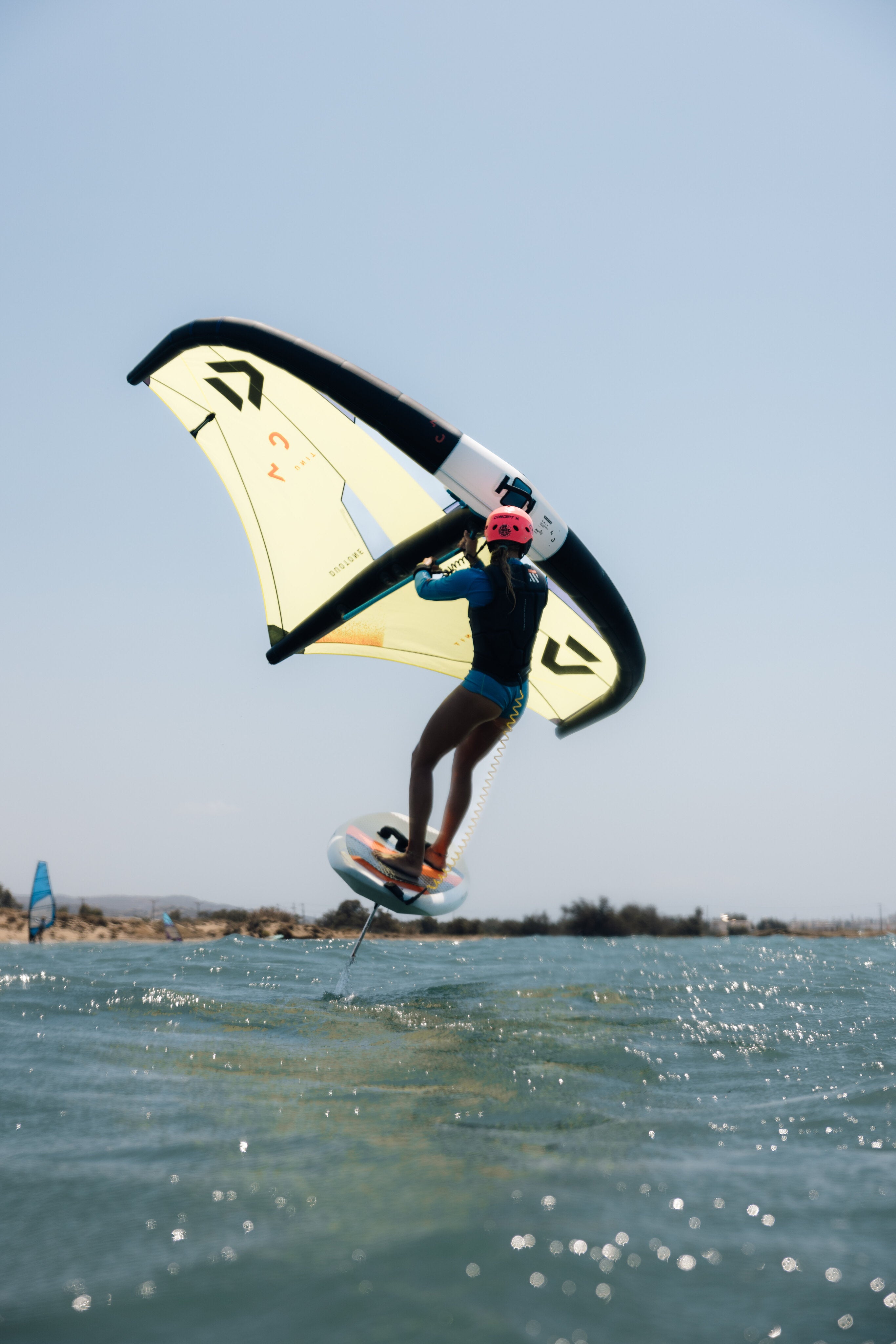 wingfoiler, roze helm, gele wing, board uit water, blauw water, blauwe lucht.