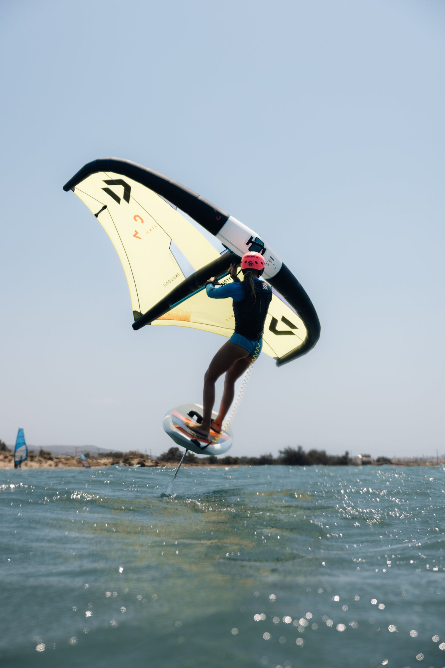 wingfoiler, roze helm, gele wing, board uit water, blauw water, blauwe lucht.