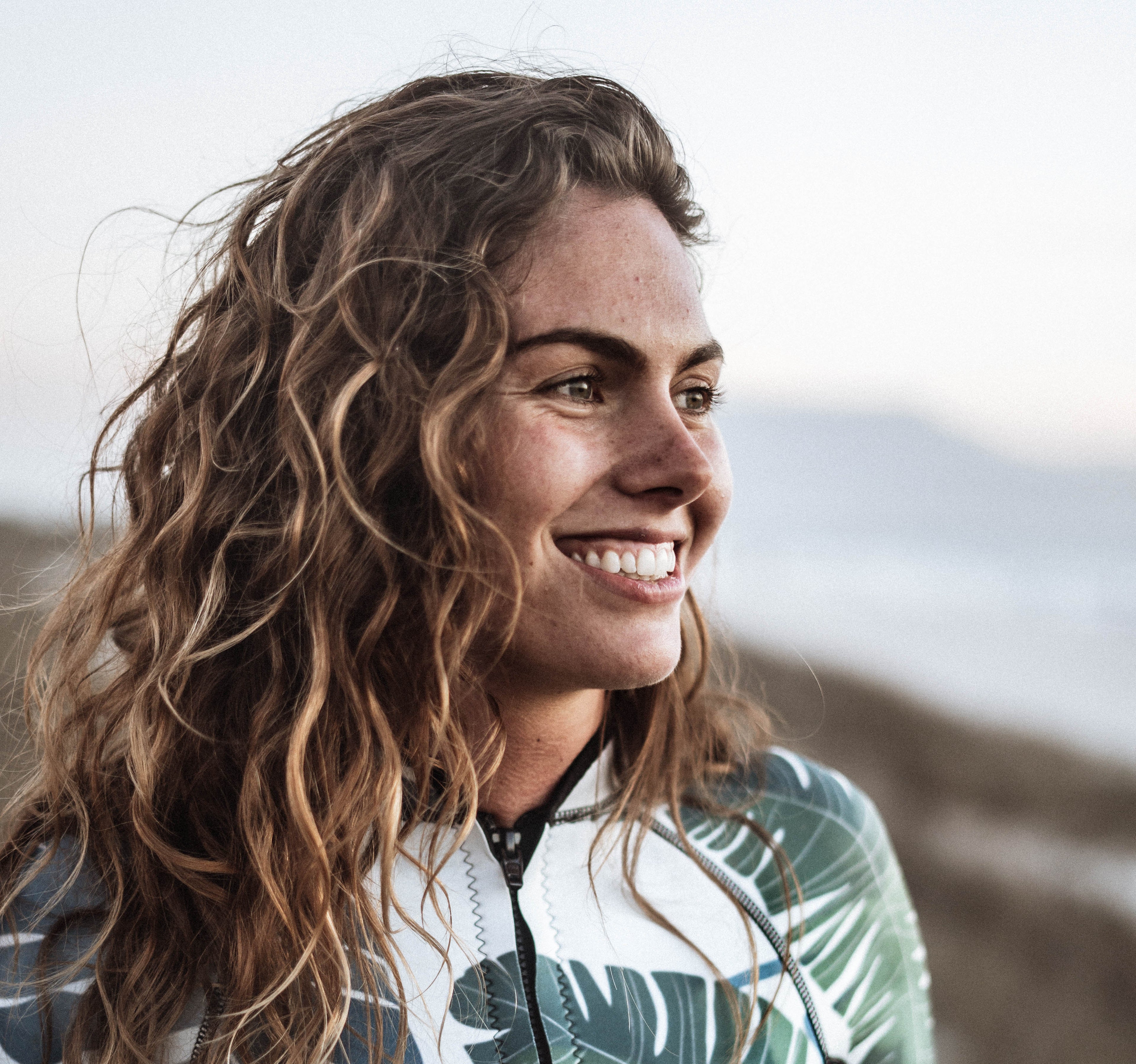 Lachende vrouw, bruin krullend haar, bruine ogen, vest met print van bladeren. 