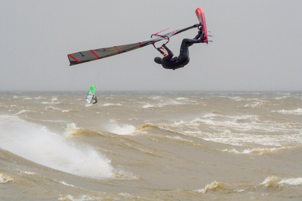 Windsurfer maakt hoge sprong, rood board, zwart met rood zeil, storm achtig weer
